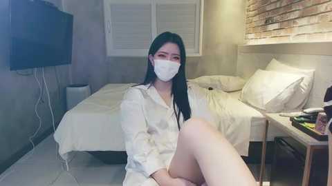 Media: Video of an Asian woman with long black hair, wearing a white mask, pajamas, and seated on a bed in a minimalist room with white walls, a TV, and a wooden nightstand.