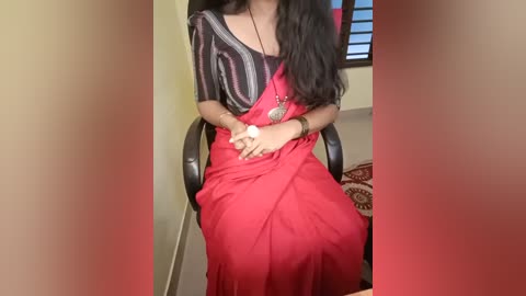 Media: Video of a woman with long black hair wearing a red saree and black blouse, seated in a chair with a window in the background, indoors.