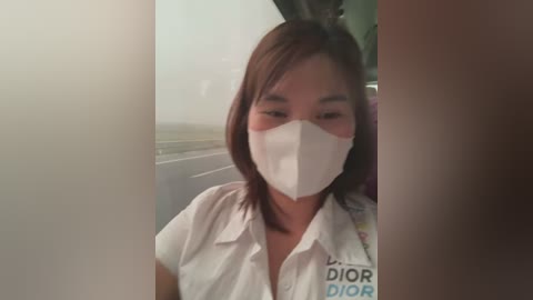 Media: Video of a young Asian woman with medium-length brown hair, wearing a white Dior blouse and a face mask, sitting in a car with a blurred outdoor scene visible through the window.