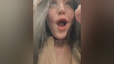 Media: Video of a fair-skinned woman with long, wavy hair, wearing round glasses, a choker, and a beige jacket, mouth open in surprise or excitement.