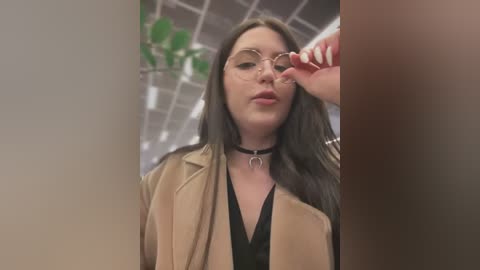 Media: Video of a young woman with long dark hair, wearing a beige blazer over a black V-neck top, and a black choker with a crescent moon pendant. She adjusts her round glasses. Background includes green plants and a ceiling with fluorescent lights.