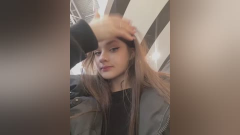 Media: A video of a young woman with long, straight brown hair, wearing a black top and a grey jacket, sitting in a car with her hand on her forehead.