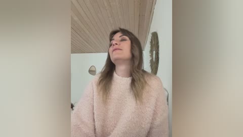 Media: Video of a fair-skinned woman with long, light brown hair, wearing a light pink fuzzy sweater, standing in a minimalist room with light wood paneling and a round mirror.