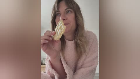 Media: A video of a middle-aged woman with light skin and long, wavy blonde hair, wearing a pink fuzzy robe, eating a cracker. Background shows a blurred, beige wall.