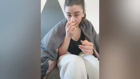 Media: A candid video of a woman in a gray cardigan and white pants, sitting on a couch with her hands to her face, expressing concern or sadness.