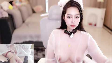 Media: Video of an Asian woman with long black hair, wearing a white maid outfit with cat ears, a collar, and a sheer top, kneeling on a bed in a modern living room.