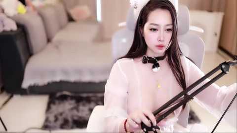 Media: Video of a young Asian woman with long black hair, wearing a white, sheer dress with a black choker and a white cat ear headband, sitting at a desk with a microphone and headset.