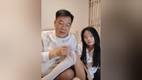 Media: A video of an elderly East Asian man and woman in a living room. The man, wearing a white sweater, sits on a couch, holding a small plastic bottle. The woman, with long black hair, sits next to him, smiling.