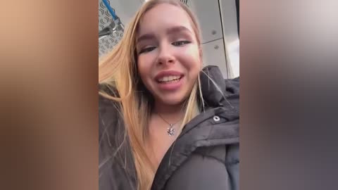 Media: A video of a young Caucasian woman with fair skin, straight blonde hair, and a smile, wearing a black puffer jacket, taken indoors with blurred background.