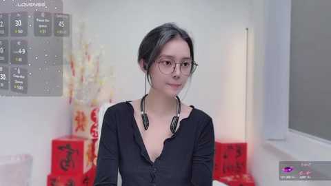 Media: Video of an Asian woman with light skin and long black hair in glasses, wearing a black top, sitting in a brightly lit room with red Chinese New Year decorations.