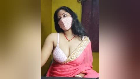 Media: Video of an Indian woman with medium skin tone and long black hair, wearing a pink mask, a white bra with polka dots, and a red sari with golden embroidery, in a yellow and purple room.