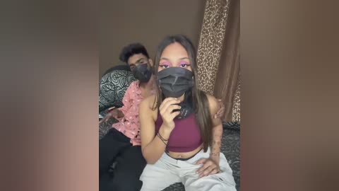 Media: A video of two people, a young woman in a pink crop top and a man in a black mask, sitting on a couch with leopard-print pillows, both wearing casual attire.