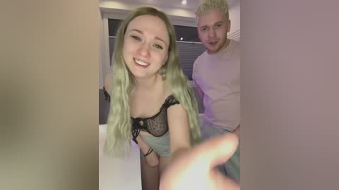 Media: Video of a smiling young woman with long, wavy, pastel green hair, wearing a black lace bra, and a man with short, platinum blonde hair, both seated indoors.