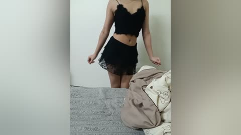 Media: Video of a slender woman with fair skin wearing black lace lingerie, standing in a messy bedroom with grey and beige bedding.