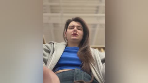 Media: Video of a young woman with long brown hair, wearing a blue crop top and denim shorts, sitting on a white bed with a beige blanket, looking down.