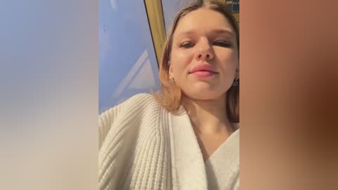 Media: A video of a Caucasian woman with fair skin and brown hair, wearing a white, textured robe, standing in front of a window with a blue sky and golden frame.