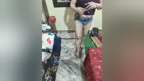 Media: A video of a woman in a small room with a cluttered floor, holding a smartphone. She wears a purple crop top and short denim shorts.