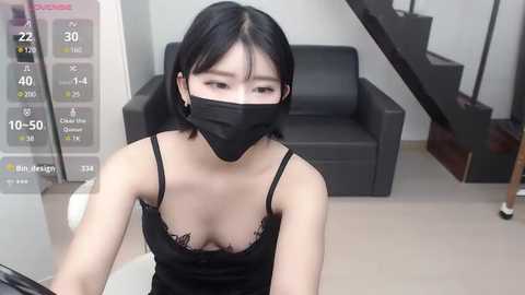 Media: Video of an Asian woman with fair skin, straight black hair, and a black face mask, wearing a black lace-trimmed tank top revealing cleavage, sitting in a modern living room with a black leather couch and a spiral staircase.