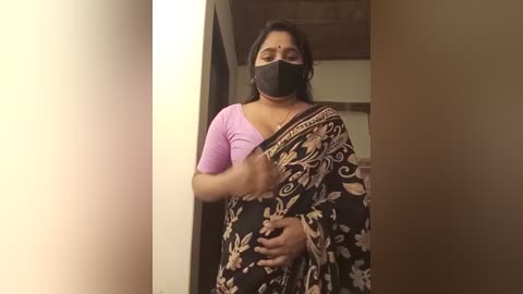 Media: Video of a South Asian woman with medium skin tone, wearing a black sari with floral patterns, pink top, black face mask, and black hair, standing indoors, against a beige wall.