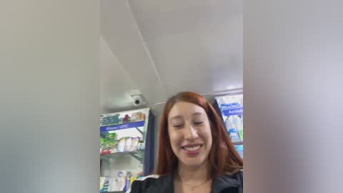 Media: Video of a smiling Asian woman with long, straight red hair, wearing a black jacket, standing in a well-lit pharmacy aisle with shelves filled with various products.