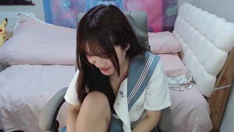 Media: Video of a young Asian woman with long black hair and fair skin, wearing a white sailor school uniform, sitting on a bed in a bedroom with pink and white bedding.