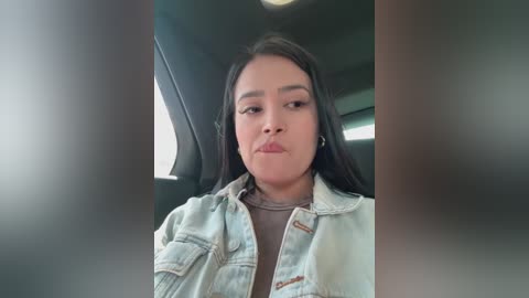 Media: A video of a young Latina woman with long black hair, fair skin, and a pouty face, wearing a denim jacket over a brown top, sitting in a car.