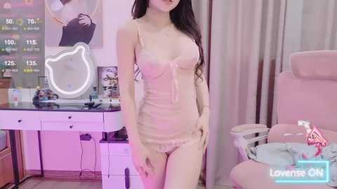 Media: Video of a slim, light-skinned woman with long dark hair, wearing a pale pink lace lingerie set, standing in a pink-themed room with a desk, chair, and makeup mirror.