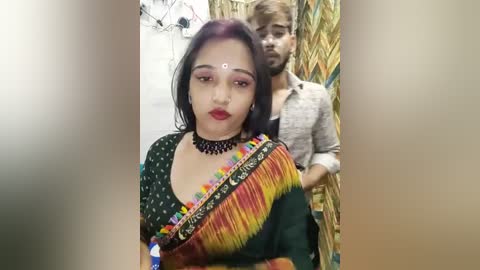 Media: Video of a young woman with dark hair and red lipstick, wearing a traditional green sari with colorful border, against a rustic background.