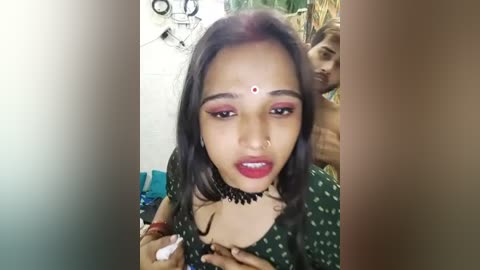 Media: Video of a young South Asian woman with medium brown skin, wearing a black polka-dot dress, red lipstick, and a red bindi, holding a white substance in her hands.