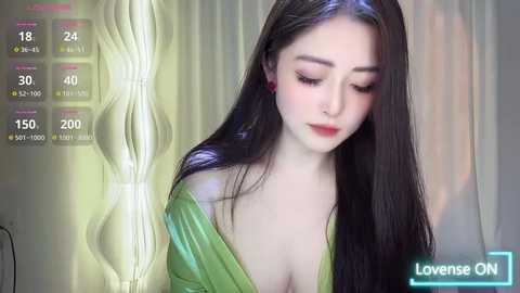 Media: Video of an Asian woman with long black hair, wearing a green off-shoulder dress, looking down. Background features a digital interface with stats and a \"Lowense ON\" button.