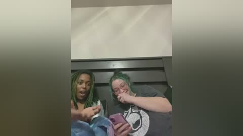 Media: A video of two women with green hair, one wearing a tank top and jeans, the other a gray T-shirt, sitting on a bench in a dimly lit, beige-walled room, laughing and holding a phone.