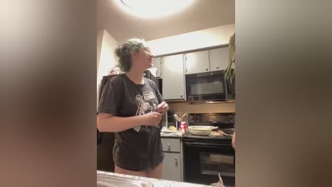 Media: A video captures a woman with short, green hair wearing a black graphic t-shirt, standing in a modern kitchen with white cabinets and black appliances.