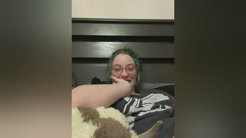 Media: Video of a young woman with light green hair, glasses, and a gray shirt, lying in bed, holding a stuffed animal, against a black and white striped headboard.