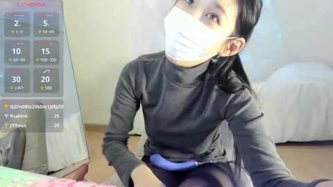 Media: Video of an Asian woman with long black hair, wearing a face mask, grey turtleneck, and blue skirt, sitting on a wooden floor, looking at a calendar on a mirror.