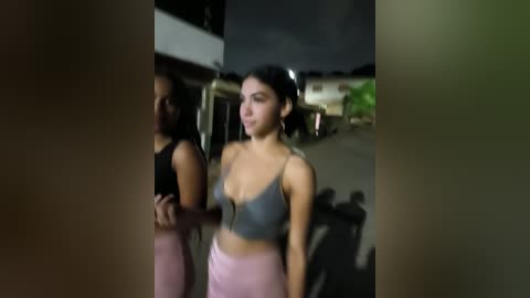 Media: Video of two women, one in a black tank top, the other in a gray crop top and pink pants, standing at night near a building.