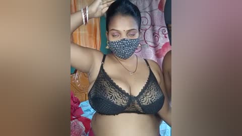 Media: Video of a South Asian woman with medium-dark skin, dark hair tied back, wearing a black lace bra, face mask, and bracelets, posing in a bedroom with colorful, patterned curtains and a bed in the background.