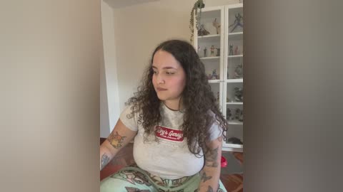 Media: Video of a curly-haired woman in a white t-shirt with a red logo, wearing floral pants, sitting indoors with a white bookshelf in the background.