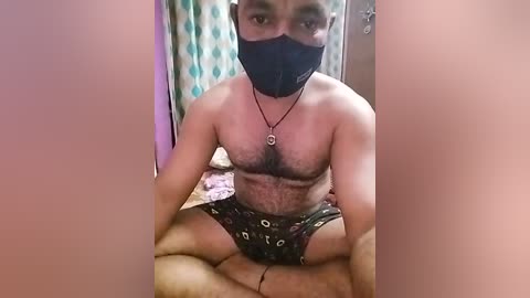 Media: Video of a shirtless South Asian man with a dark beard and black mask, sitting cross-legged on a bed in a dimly lit room, wearing black shorts with white patterns.