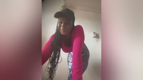 Media: Video of a young Black woman with long braids, wearing a pink long-sleeve shirt, floral leggings, and a black cap, leaning forward against a beige wall in a dimly lit room.