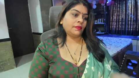 Media: Video of a middle-aged woman with dark skin, wearing a green and red checkered blouse, gold necklace, and a green saree with white floral design. She sits in a modern office with a gray chair, wooden desk, and a patterned rug in the background.