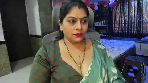 Media: Video of a middle-aged South Asian woman with medium skin tone, wearing a green sari with white floral motifs, seated indoors. Background shows a bed, a wooden cabinet, and a dark curtain.