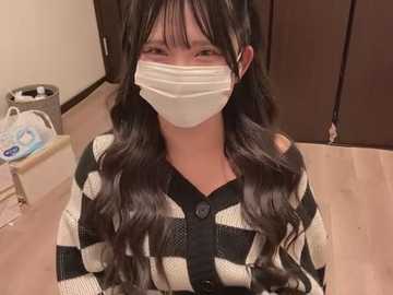 Media: Video of an Asian woman with long, wavy black hair and a white face mask, wearing a black and white plaid sweater, seated at a wooden table in a room with brown cabinets and a trash bin.