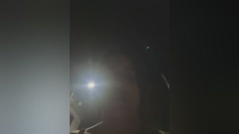 Media: A video with a dark, blurry background features a person holding a smartphone with a bright, blinding flash, creating a lens flare effect. The person\u2019s face is mostly obscured by the light, suggesting a selfie or close-up shot.