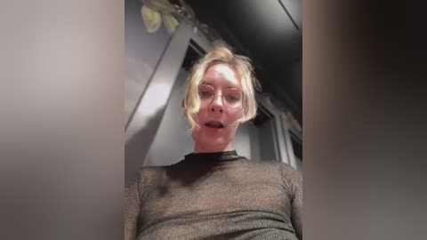 Media: Video of a blonde woman with short hair, wearing a sheer black top, standing indoors with blurred background.