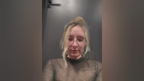 Media: A video of a fair-skinned woman with blonde hair tied back, wearing glasses and a sheer, long-sleeved top, standing in a dimly lit, plain hallway.