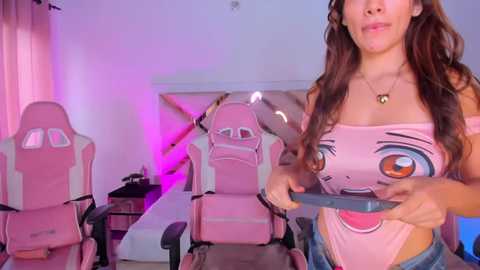 Media: Video of a light-skinned woman with long brown hair, wearing a pink strapless top with a cartoonish face, holding a controller. She stands in a room with pink gaming chairs and a staircase.