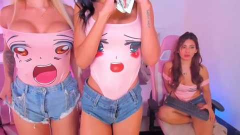 Media: Video of three young women in a gaming room; two wear tight, pink bodysuits with cartoon faces, denim shorts; one sits in a pink gaming chair, wearing a pink crop top and shorts.