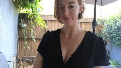 Media: Video of a light-skinned woman with short brown hair, wearing a black V-neck top, smiling in a backyard. Background includes a wooden fence, greenery, and a beige patio umbrella.