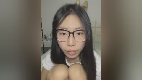 Media: Video of an Asian woman with long black hair, wearing glasses and a white shirt, sitting with knees up, in a home setting with a blurred background.