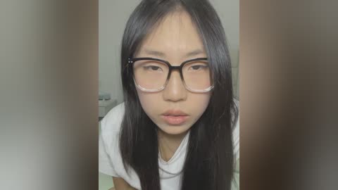 Media: A close-up video of an Asian woman with long black hair, wearing large, rectangular glasses and a white t-shirt, leaning forward with a neutral expression. The background is blurred, featuring beige walls.